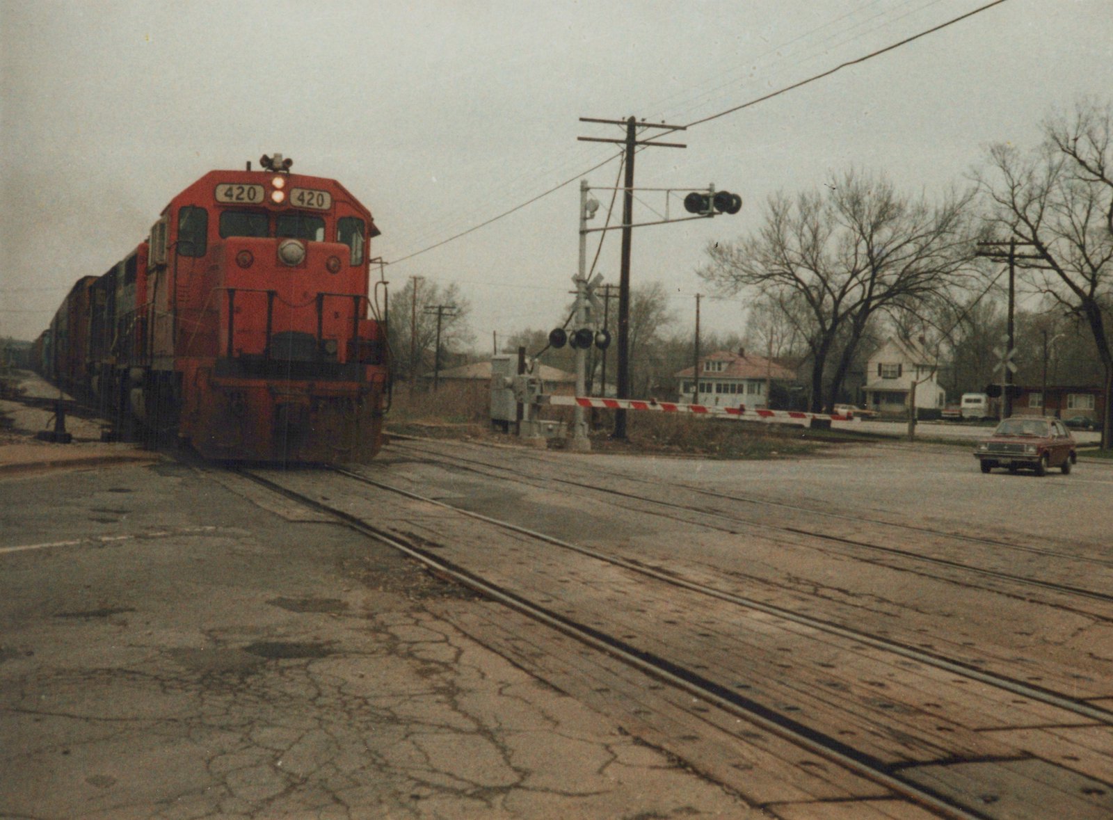 A red Grand Trunk Western diesel locomotive numbered 420 is crossing a street at a railroad crossing, with a red car waiting on the side and houses in the background.