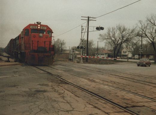 A red Grand Trunk Western diesel locomotive numbered 420 is crossing a street at a railroad crossing, with a red car waiting on the side and houses in the background.
