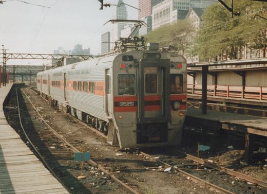 A silver South Shore Line Highliner train with red and black stripes, numbered 5, sits on an elevated track next to a wooden platform, with Chicago skyscrapers in the background.