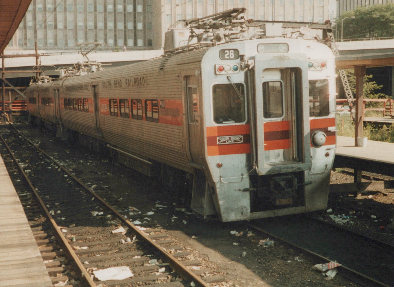 A silver and orange double-decker electric train, marked 'South Shore Line' and numbered 26, is stopped at an elevated station platform.
