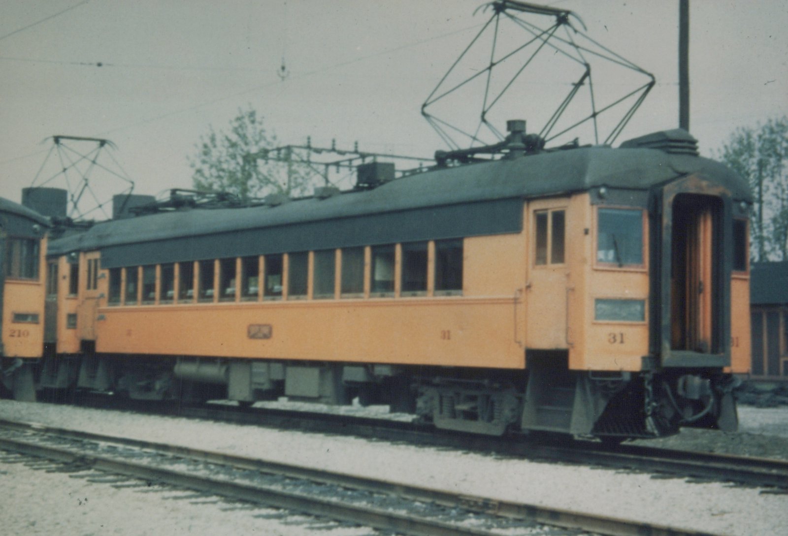 Side view of two orange and green interurban cars, numbered 31 and 210, on tracks in a yard under an overcast sky.