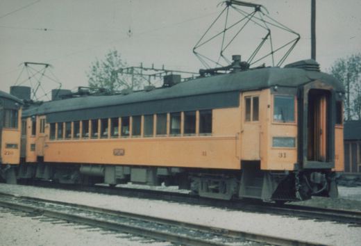 Side view of two orange and green interurban cars, numbered 31 and 210, on tracks in a yard under an overcast sky.
