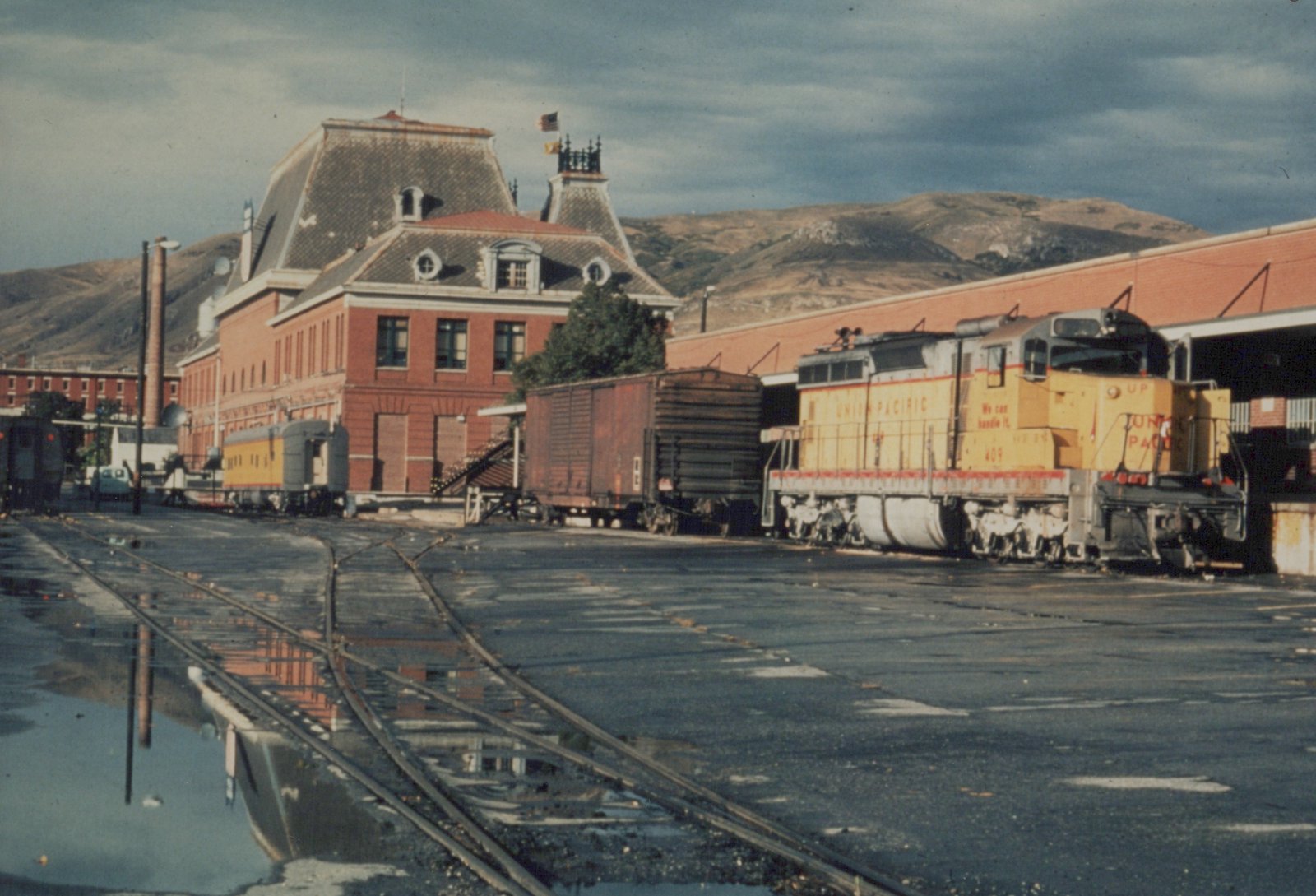A yellow Union Pacific SD24 locomotive, number 409, is parked on tracks at a train station with a red brick building and a mountain in the background.