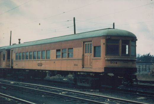 An orange and green interurban car numbered 504 from the Chicago South Shore and South Bend Railroad, viewed from a three-quarter front angle, stopped on tracks with overhead wires visible.
