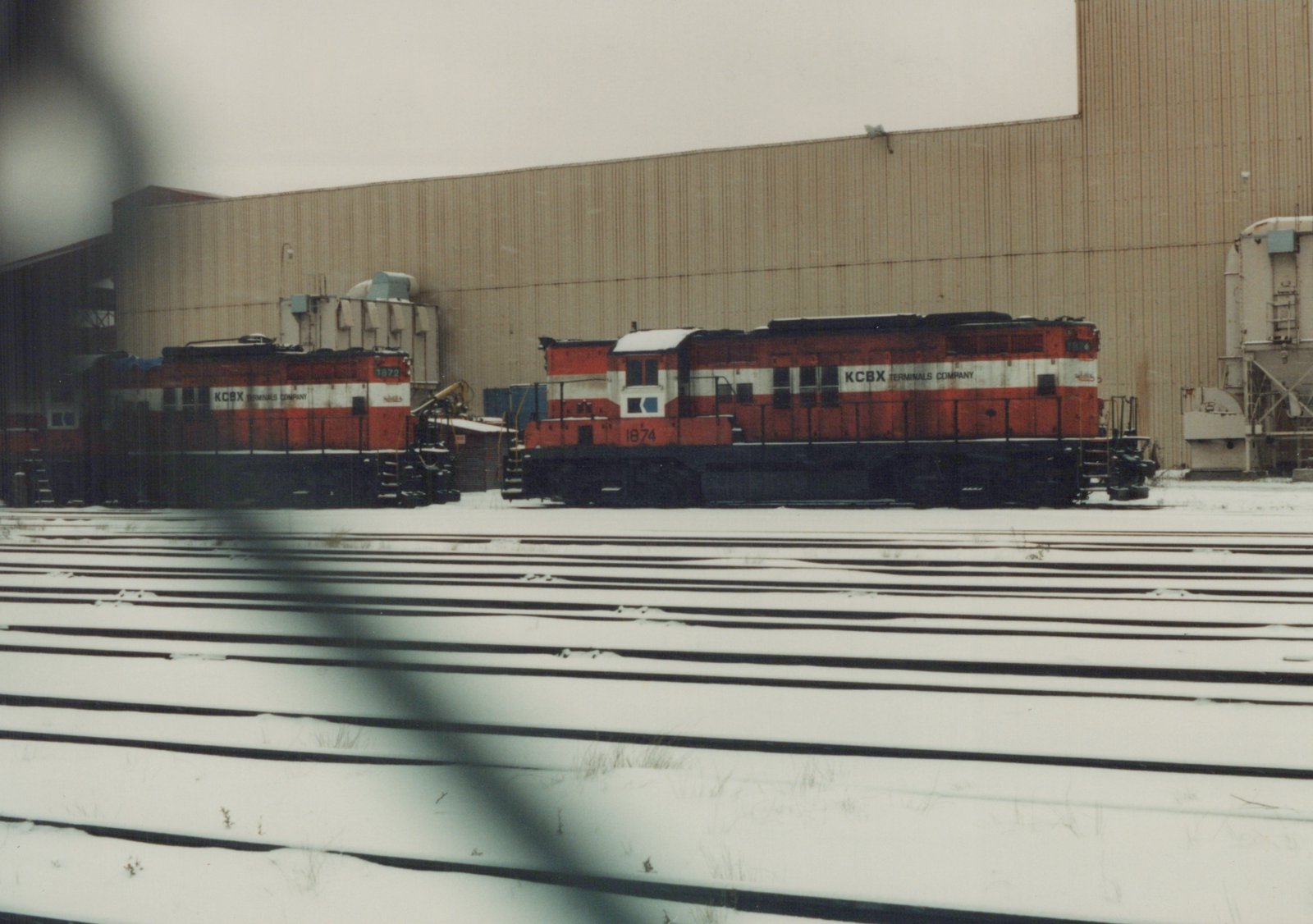 Two red and white diesel locomotives with KCBX lettering are parked on parallel tracks in a snowy yard with a large industrial building behind them.