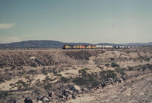 A long freight train with yellow and blue locomotives moves across a barren, rocky desert under a clear blue sky with distant mountains.