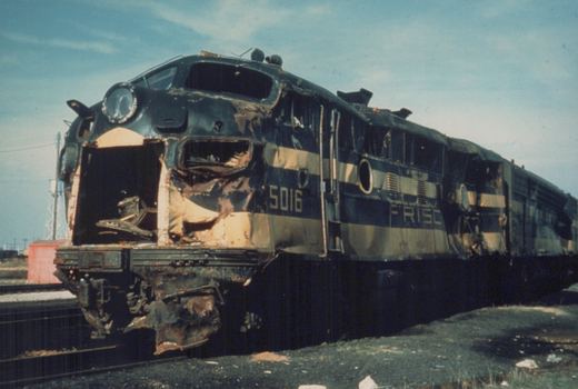 A wrecked Frisco diesel locomotive, number 5016, with a destroyed front end and burned paint, rests on a railroad track in a yard under a clear sky.