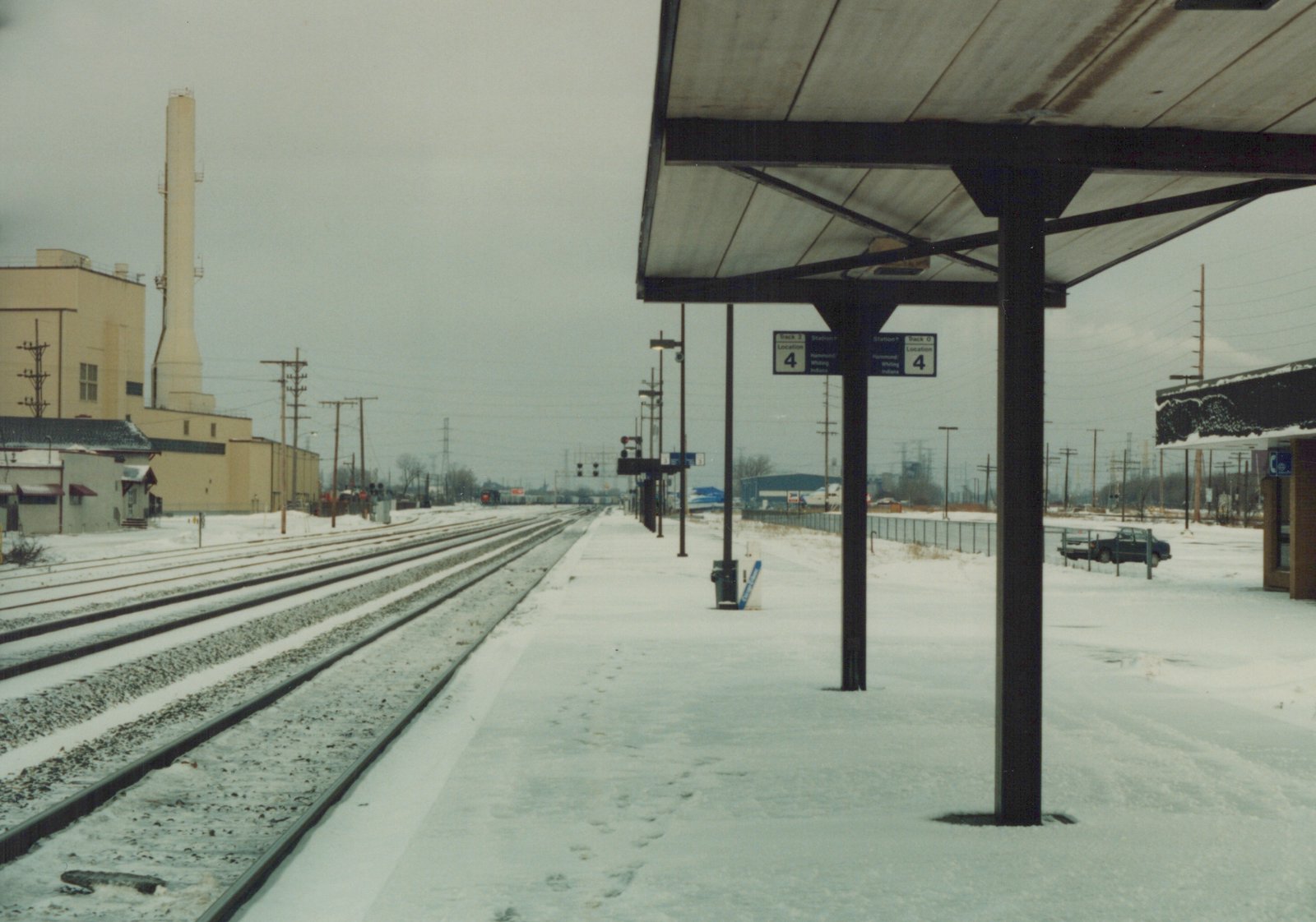 A snow-covered train station platform with signs for Track 2 and Track 0 at the Hammond-Whiting, Indiana station, with multiple tracks and a distant train visible.
