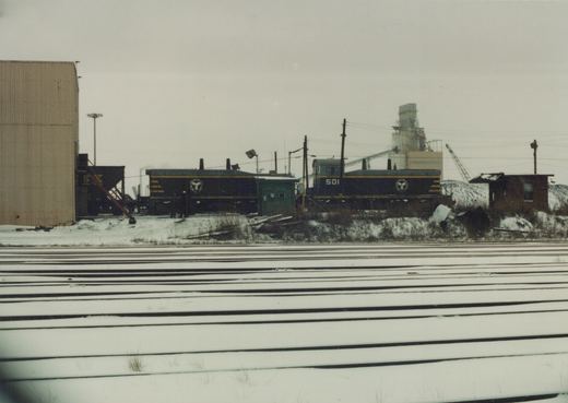 A black and yellow Soo Line locomotive numbered 501 is parked in a snowy rail yard with multiple tracks and industrial buildings in the background.