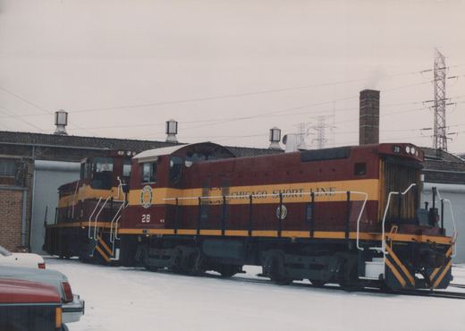 Two maroon and yellow Chicago Short Line Railway diesel locomotives stand in a snow-covered yard with industrial buildings in the background.