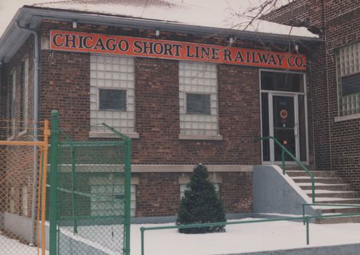 A snow-covered brick building with a large sign reading 'CHICAGO SHORT LINE RAILWAY CO.', a green metal fence, and a small evergreen tree in front.