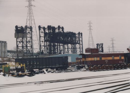 Snowy rail yard scene featuring a maroon and yellow caboose and black freight cars, with industrial steel towers and power lines in the background.