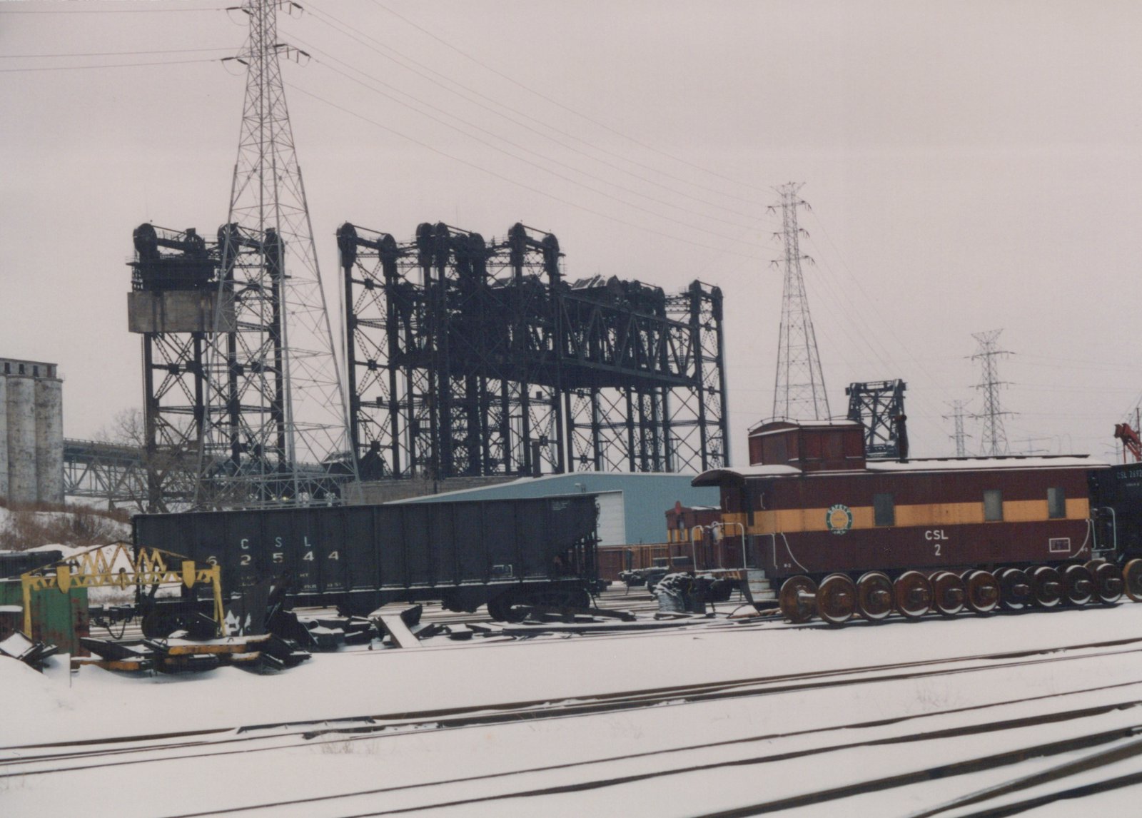 Snowy rail yard scene featuring a maroon and yellow caboose and black freight cars, with industrial steel towers and power lines in the background.