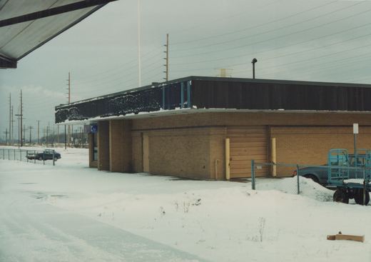 A low, brick building with a dark roof covered in snow, surrounded by a snowdrift and fenced parking area with two vehicles, under a grey sky with overhead power lines.