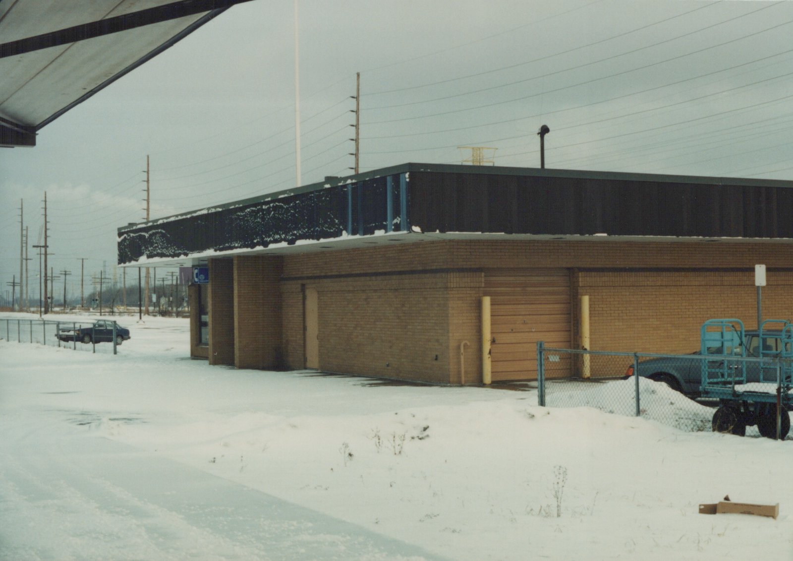 A low, brick building with a dark roof covered in snow, surrounded by a snowdrift and fenced parking area with two vehicles, under a grey sky with overhead power lines.