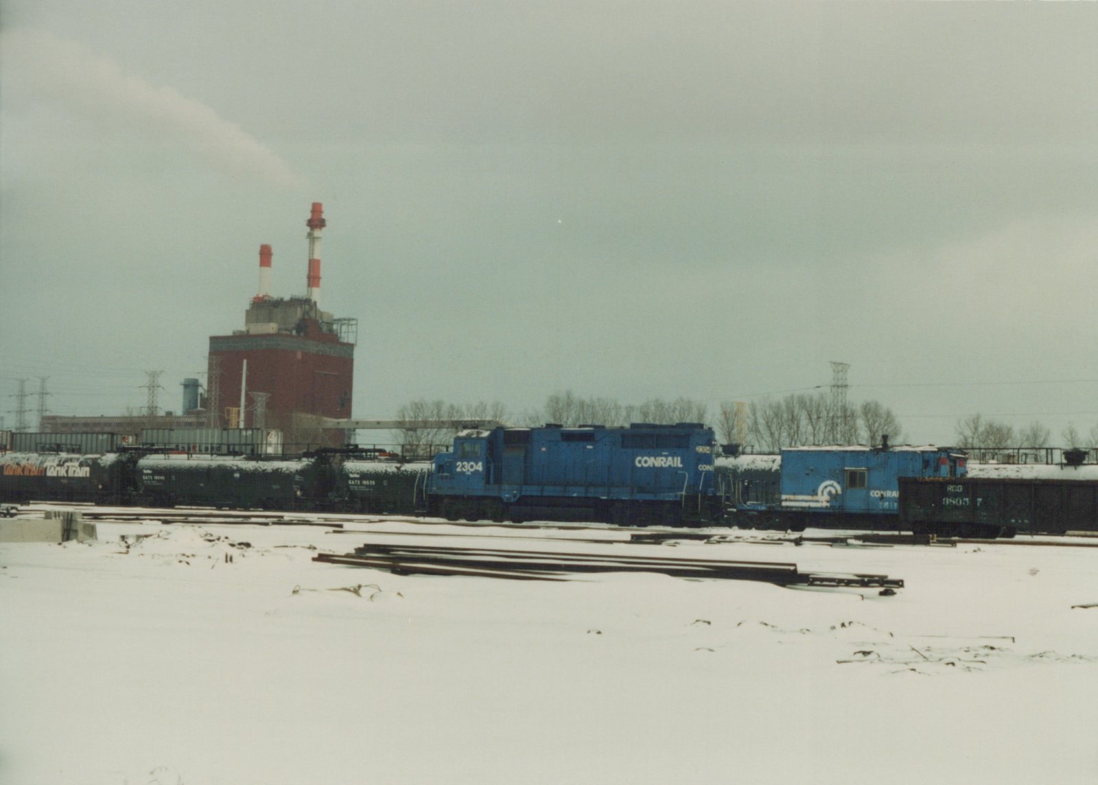 Side view of a blue Conrail diesel locomotive numbered 2304 and a blue RDG caboose numbered 8805 7, in a snow-covered rail yard with industrial buildings and bare trees in the background.
