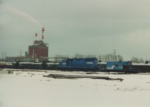 Side view of a blue Conrail diesel locomotive numbered 2304 and a blue RDG caboose numbered 8805 7, in a snow-covered rail yard with industrial buildings and bare trees in the background.