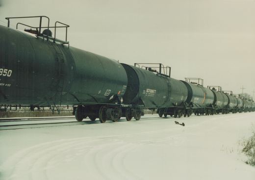 A long line of black tank cars on railroad tracks in a snowy, rural landscape. The cars are marked with ACFX reporting marks and numbers including 850 and 89677.