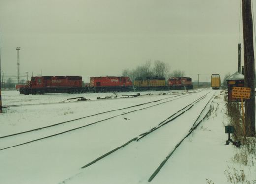 A winter scene in a railroad yard with several locomotives parked on snow-covered tracks, including Canadian Pacific red engines and a yellow GATX engine, with a sign reading 'ENGINES ARE NOT ALLOWED ON SCALE WEIGH-RAILS'.