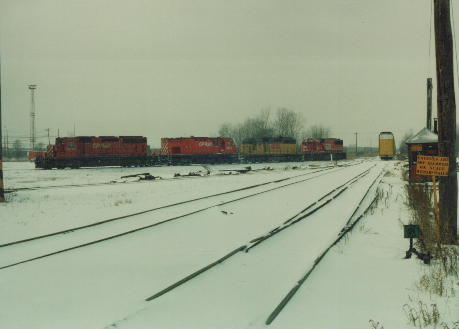 A winter scene in a railroad yard with several locomotives parked on snow-covered tracks, including Canadian Pacific red engines and a yellow GATX engine, with a sign reading 'ENGINES ARE NOT ALLOWED ON SCALE WEIGH-RAILS'.
