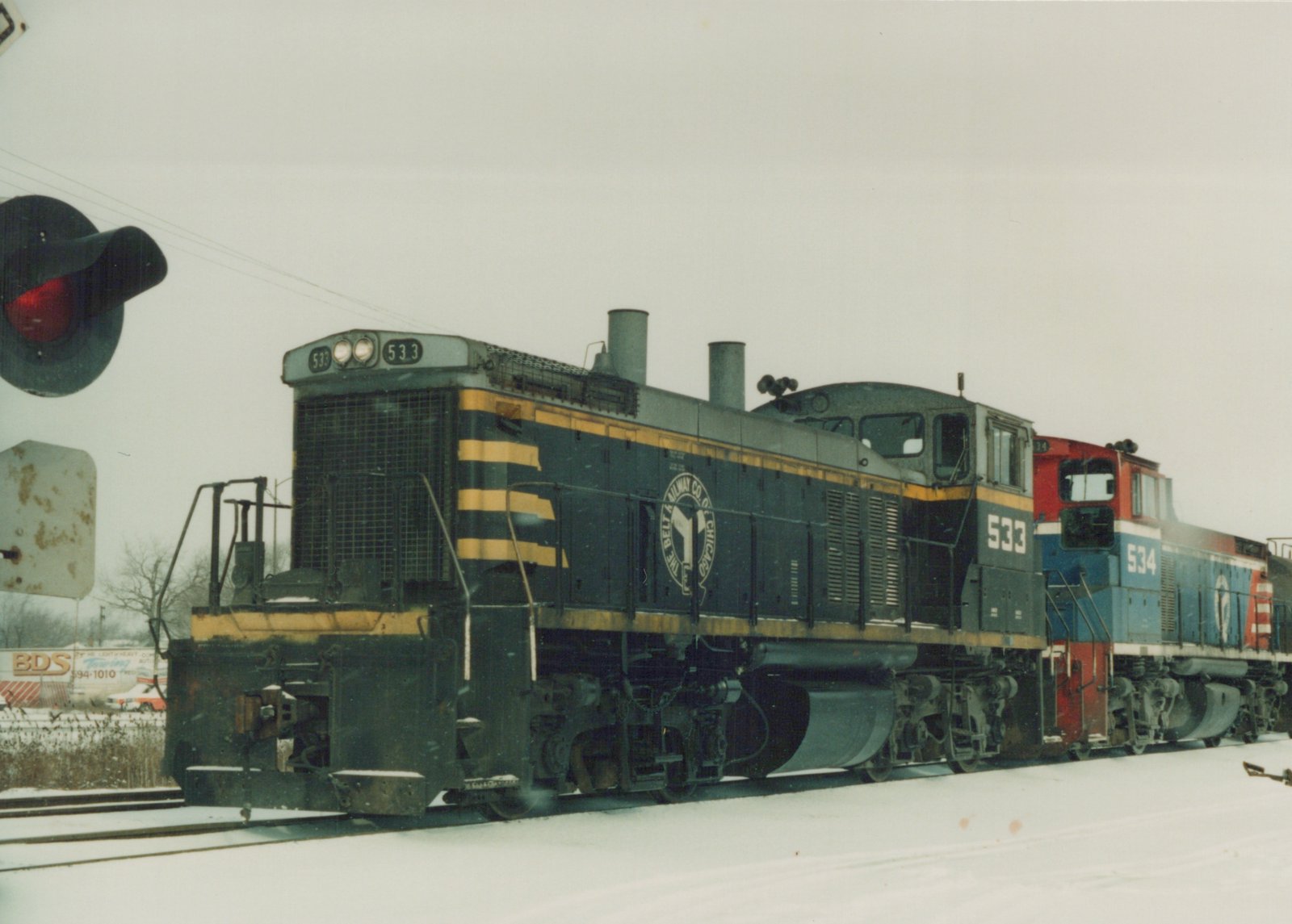 Two diesel locomotives, one black and yellow and the other red and blue, are coupled together on snow-covered tracks next to a railroad crossing signal.