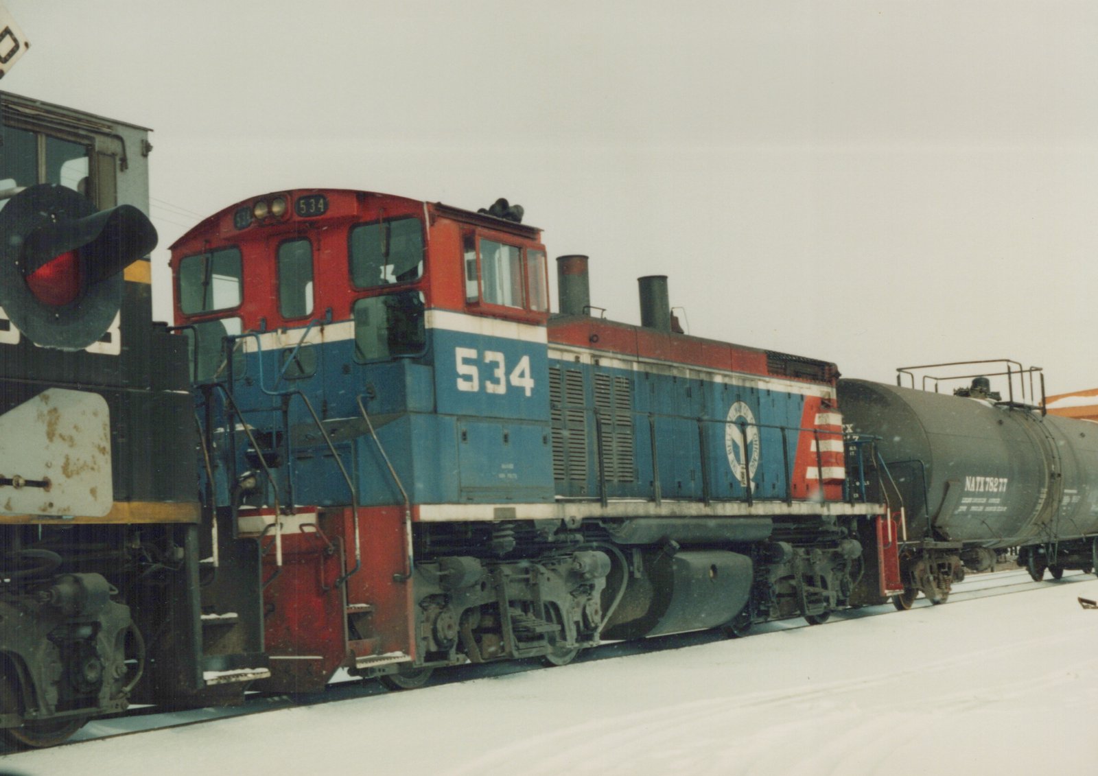 A red, white, and blue diesel locomotive numbered 534 sits beside a black tank car on a snowy track in an open yard under an overcast sky.