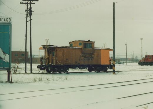 A side view of an orange caboose with the number 01512 and 'Rio Grande' lettering, parked on snowy tracks, with a billboard reading 'CHICAGO' and a red locomotive visible in the background.