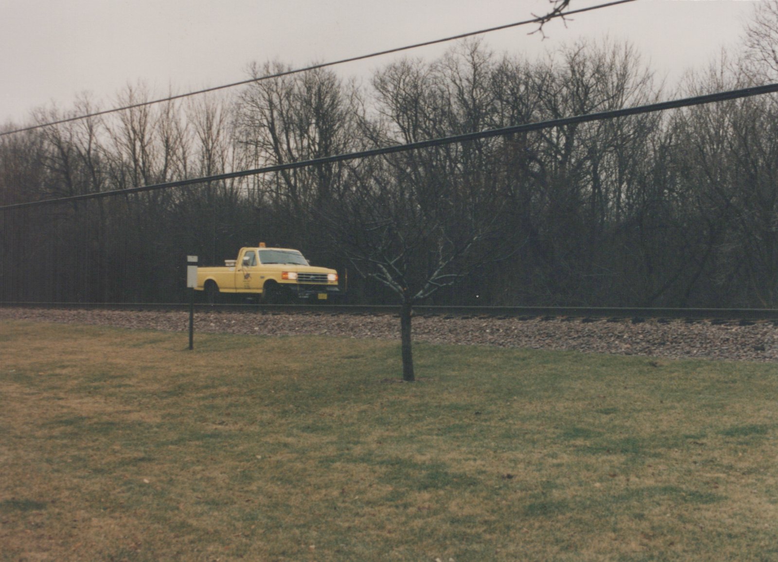 A yellow pickup truck parked on a gravel bed next to railroad tracks, surrounded by bare trees and grass under a gray sky.