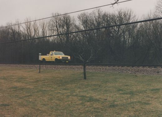 A yellow pickup truck parked on a gravel bed next to railroad tracks, surrounded by bare trees and grass under a gray sky.