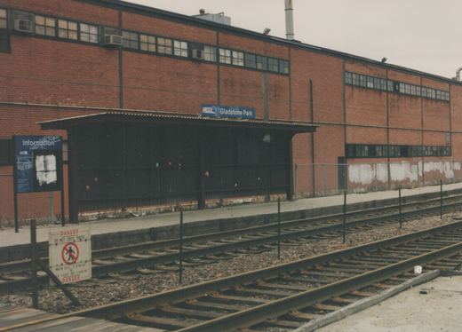 An empty train station platform with a brick building behind it. A sign above reads 'Metra Gladstone Park'. A danger sign near the tracks warns not to cross when the bell is ringing or red lights are flashing.