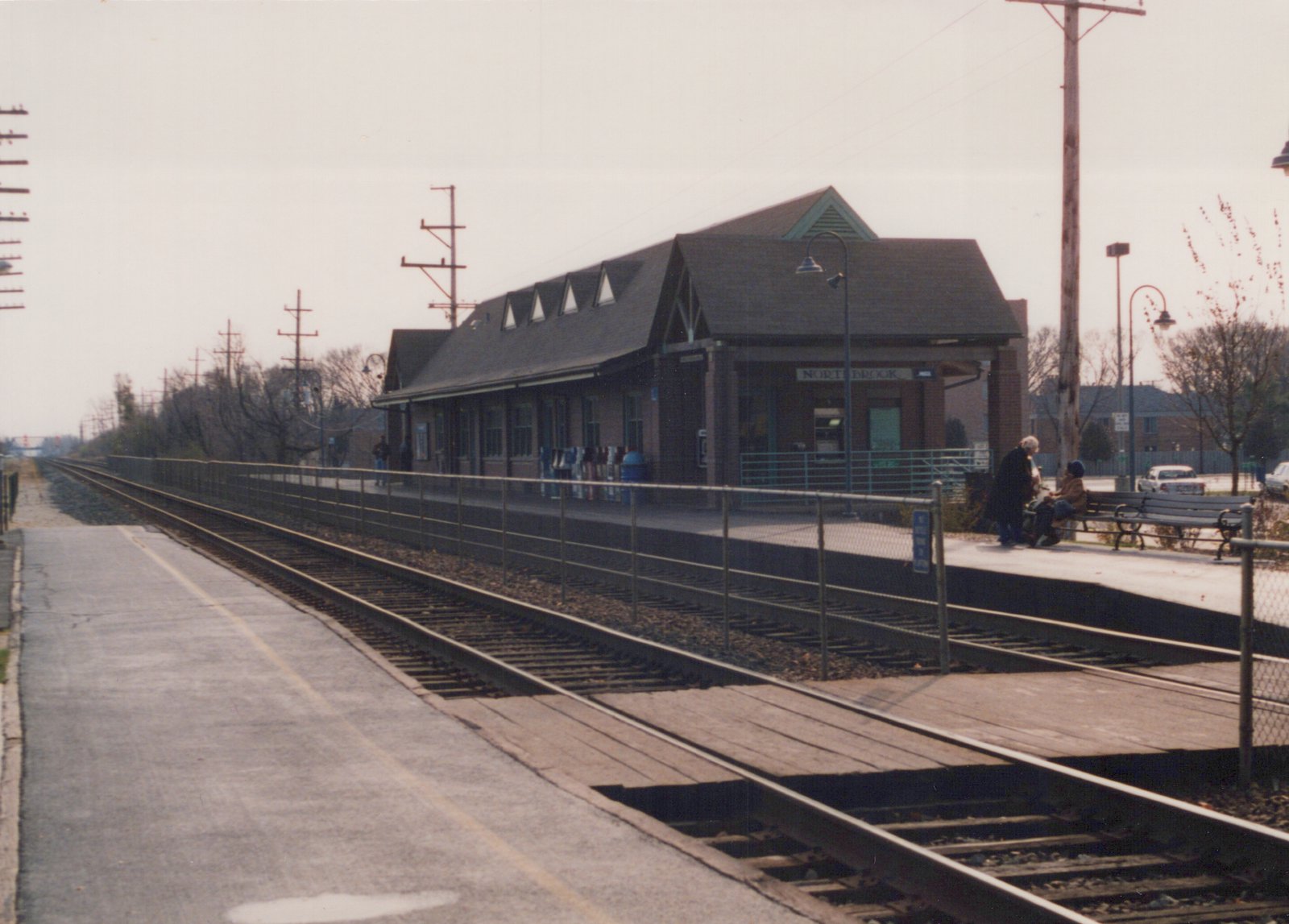 A quiet, overcast day at the Northbrook train station with no trains, two people on a bench, and a modern brick building in the background.