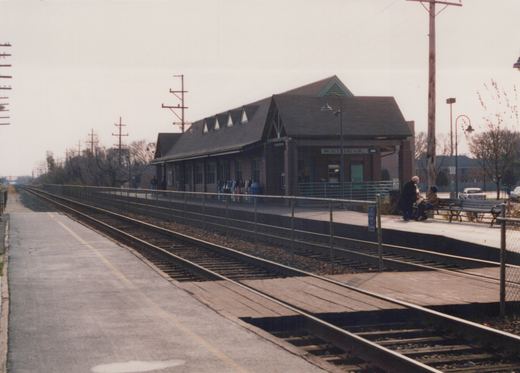 A quiet, overcast day at the Northbrook train station with no trains, two people on a bench, and a modern brick building in the background.