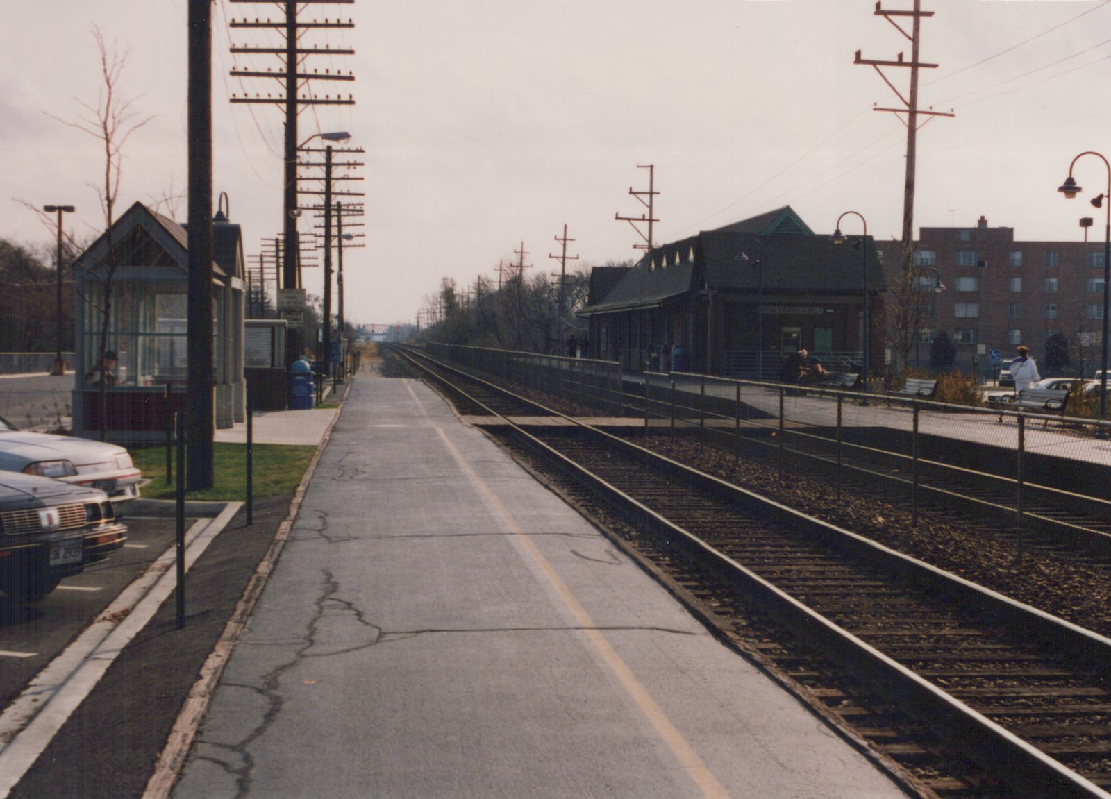 A view down a cracked concrete platform at a commuter rail station, with two sets of tracks, a small station building, and a few people and cars visible.