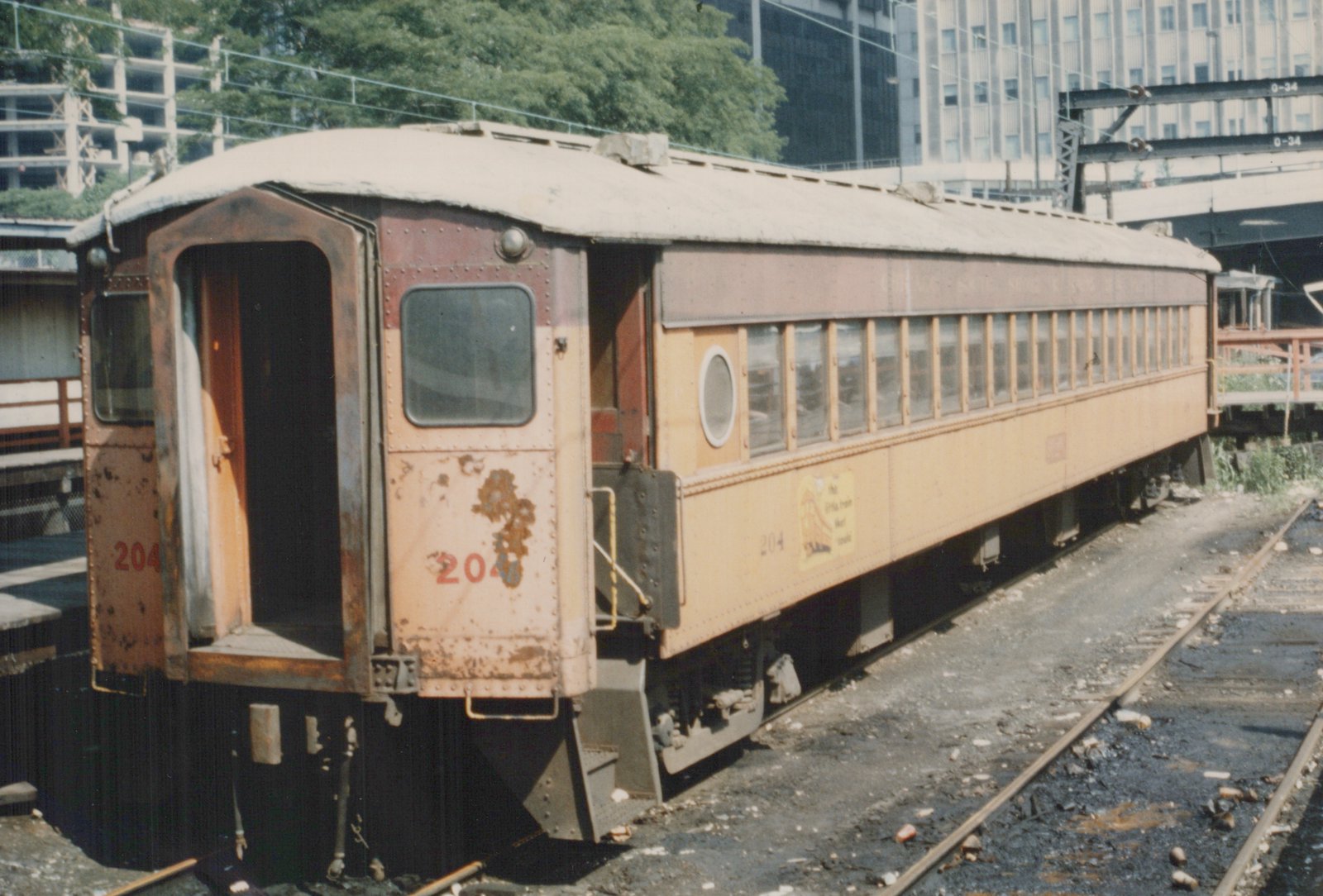 A side-view of a rusty, yellow and maroon interurban railcar numbered 204, parked on tracks with a city building and an elevated train structure in the background.