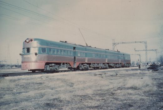 A long, streamlined electric train with a blue and red paint scheme and a silver upper body stands on railroad tracks in a snowy, open field under an overcast sky.