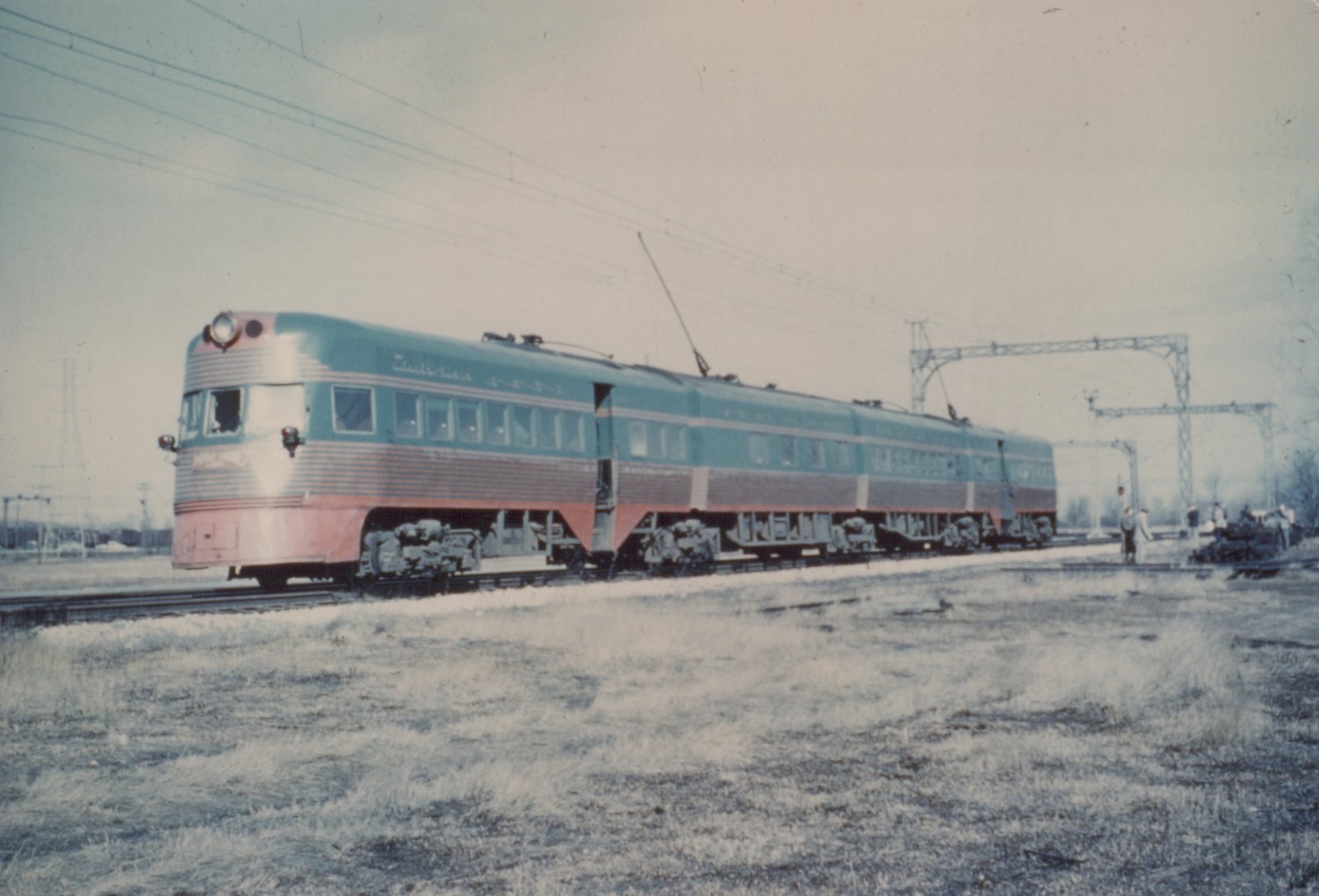 A long, streamlined electric train with a blue and red paint scheme and a silver upper body stands on railroad tracks in a snowy, open field under an overcast sky.
