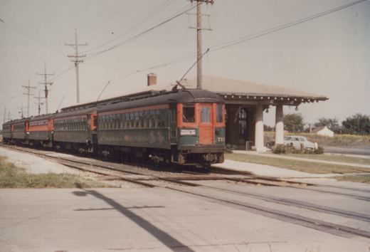 An interurban train with a green and orange car, numbered 713 and marked 'MUNDELEIN', is stopped at a station platform under overhead wires, with a 1950s car parked nearby.