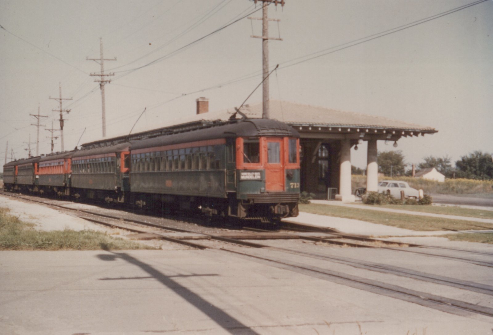 An interurban train with a green and orange car, numbered 713 and marked 'MUNDELEIN', is stopped at a station platform under overhead wires, with a 1950s car parked nearby.