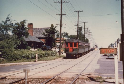 An orange and green interurban train labeled CNS&M 183 travels down a street with visible trolley wires, passing a brick station and a stop sign, with a person standing on the sidewalk and cars parked nearby.
