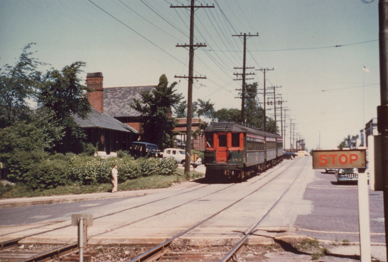 An orange and green interurban train labeled CNS&M 183 travels down a street with visible trolley wires, passing a brick station and a stop sign, with a person standing on the sidewalk and cars parked nearby.