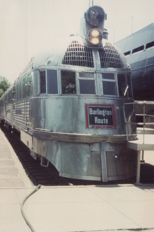 A front three-quarter view of a sleek, silver, streamlined train car with a 'Burlington Route' sign, parked at a station.