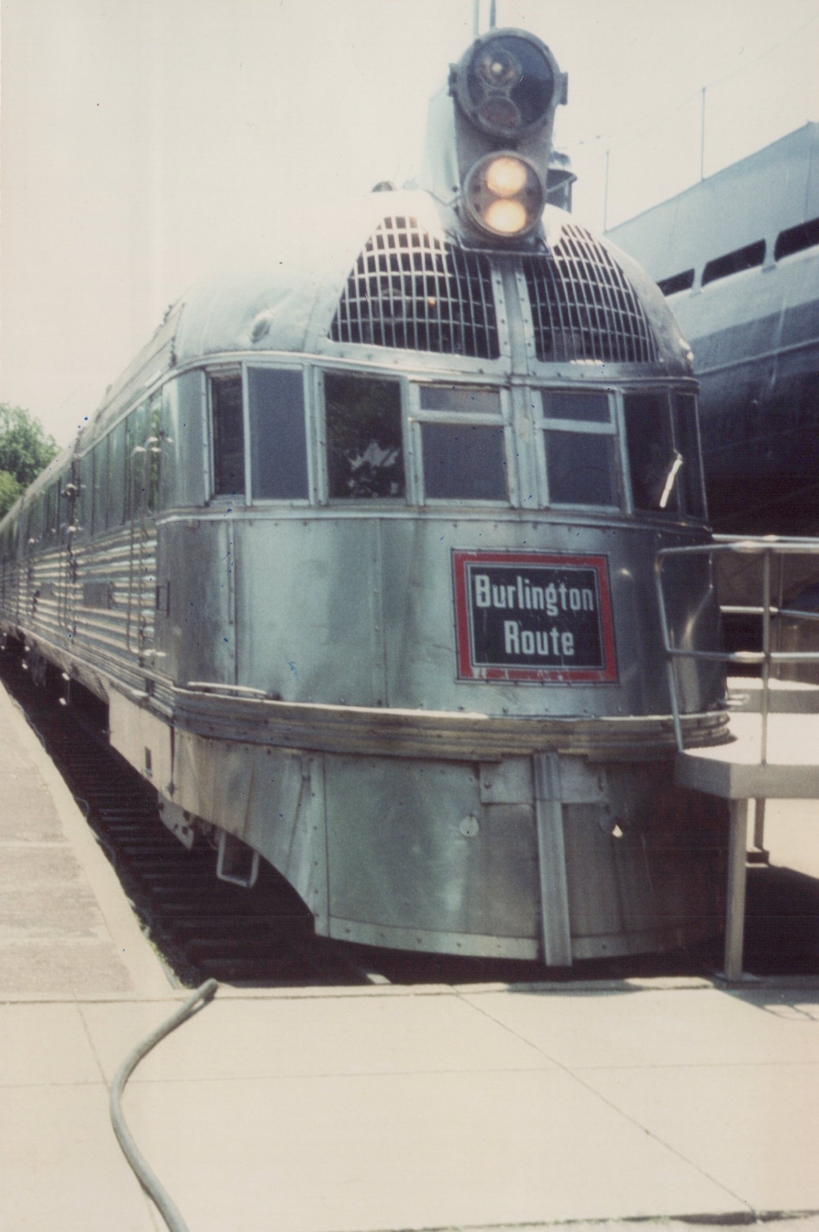 A front three-quarter view of a sleek, silver, streamlined train car with a 'Burlington Route' sign, parked at a station.