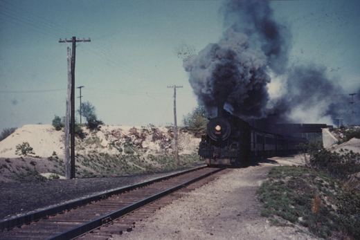 A black steam locomotive billowing thick black smoke travels on a set of tracks with embankments and utility poles on either side.