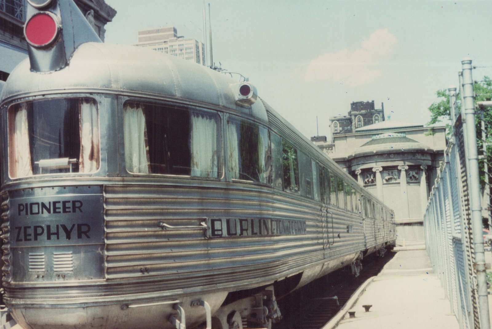 A close-up of the front and side of the Pioneer Zephyr train, showing its sleek, curved stainless steel body and 'BURLINGTON' lettering, parked beside a platform with a chain-link fence and buildings in the background.