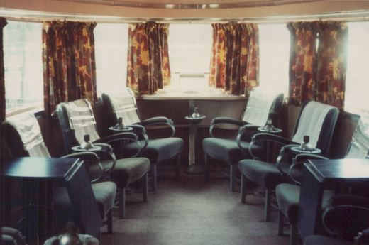 The interior of a mid-century passenger train car featuring rows of cushioned armchairs with attached ashtray tables, floral curtains over large windows, and a carpeted floor.