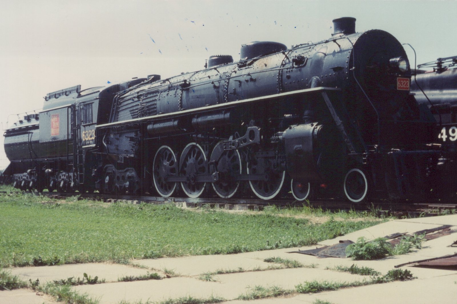 A black steam locomotive numbered 6323, labeled Grand Trunk Western, is parked on tracks next to grass and pavement, with another locomotive partially visible behind it.