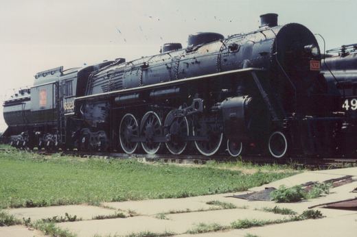 A black steam locomotive numbered 6323, labeled Grand Trunk Western, is parked on tracks next to grass and pavement, with another locomotive partially visible behind it.