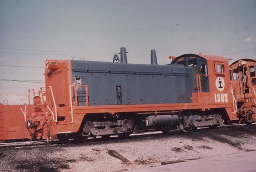 A side view of an orange and gray Illinois Central Gulf diesel locomotive, number 1303, parked on tracks in a yard with another similar locomotive behind it.
