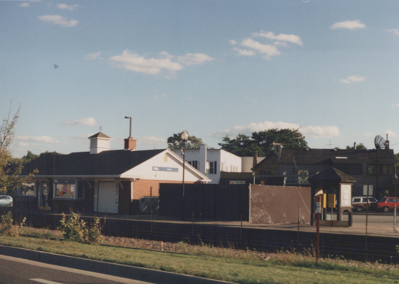 A view of the Cary train station, featuring a white station house with a blue sign, a platform shelter, and several parked cars under a blue sky with scattered clouds.