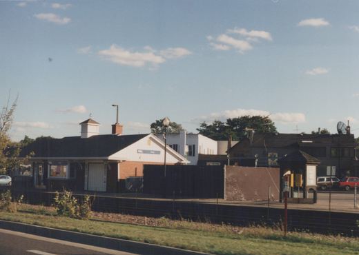 A view of the Cary train station, featuring a white station house with a blue sign, a platform shelter, and several parked cars under a blue sky with scattered clouds.